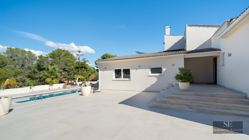 Villa blanche moderne avec piscine, grande terrasse en pierre et palmiers en pots sous un ciel bleu clair.
