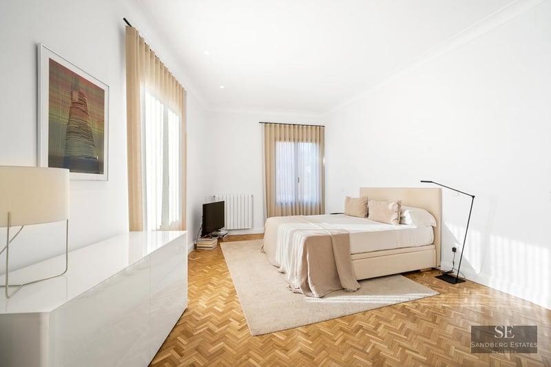 Bright bedroom with herringbone wood floor, large bed, beige curtains, and minimalist designer floor lamp.