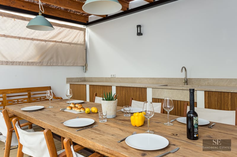 Long wooden table set for several guests on a covered terrace with an outdoor kitchen in the background.