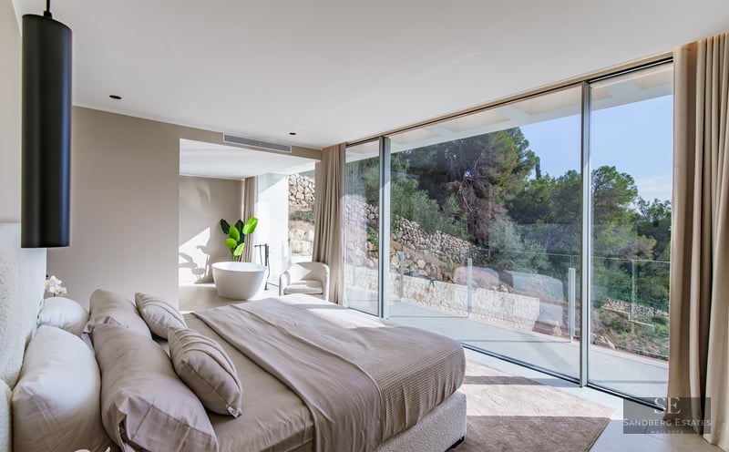 Modern bedroom with floor-to-ceiling windows, a large bed in neutral tones, and an ensuite bathtub with garden views.