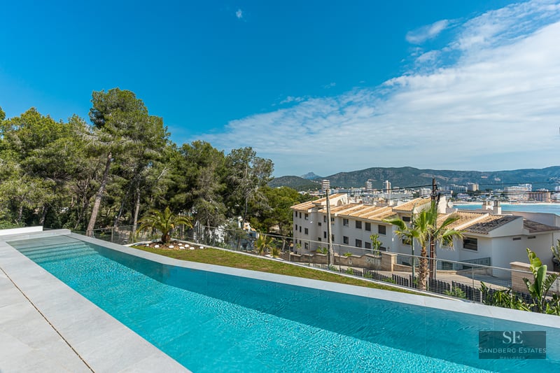 Luxury infinity swimming pool with stone tiling, overlooking a coastal town, mountains, and the sea under a clear blue sky.