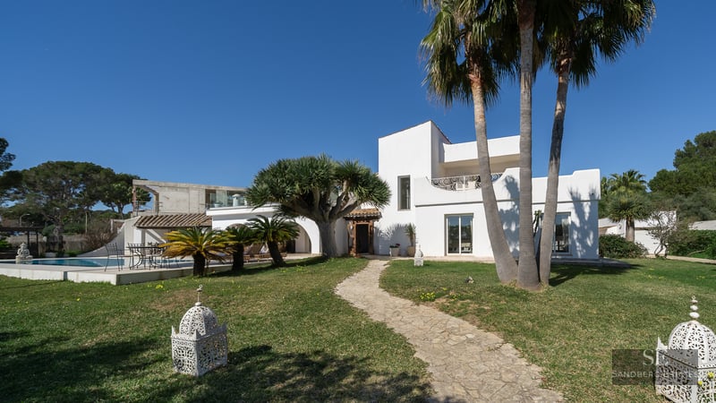 White Mediterranean villa with a winding stone path, palm trees, and a swimming pool under a bright blue sky.