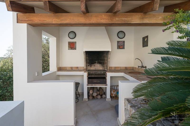 Un espace barbecue extérieur couvert avec des poutres en bois rustiques, des murs blancs, un gril en pierre et un évier.