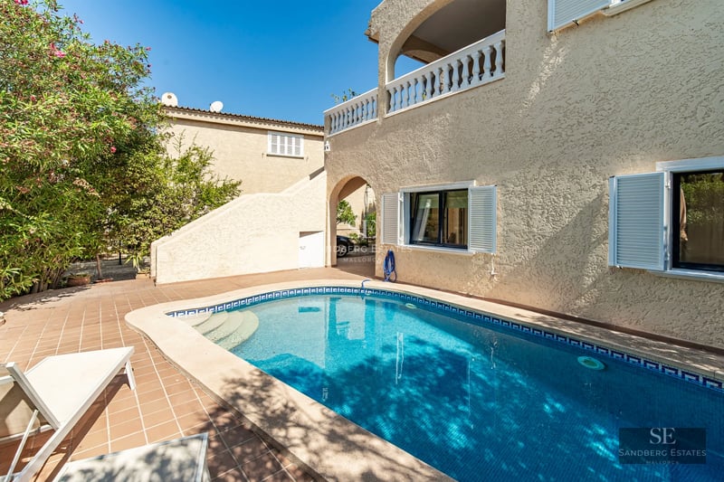 View of a pool surrounded by lawn and a modern two-story house with large windows. Natural lighting and minimalist design.