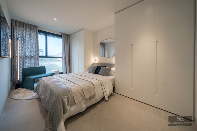 A bright modern bedroom featuring a bed with grey linens, large white wardrobes, a teal chair, and a large window.
