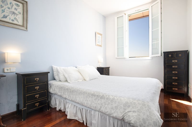 A bedroom with a white floral bed, black nightstands, dark wood floors, and an open window showing a blue sky.