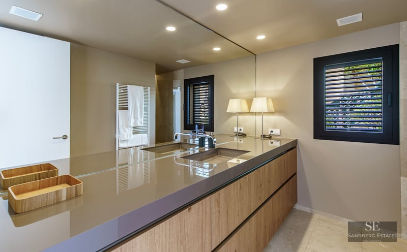 A large mirror reflects a modern wooden bathroom vanity with an integrated sink and designer table lamps.