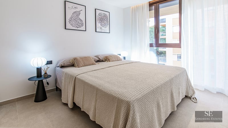 Bright bedroom with a large bed, beige waffle-knit linens, designer lamps, and a window with sheer white curtains.