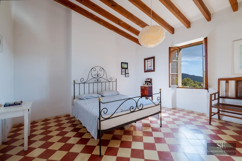 Bedroom with a black wrought iron bed, red and white checkered floor, exposed wooden beams, and a window with hill views.