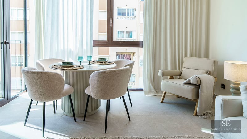 Bright dining room with round white table, four bouclé chairs, large windows with white curtains, and a beige armchair.