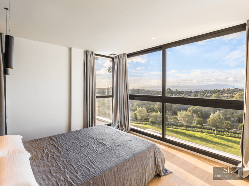 Modern bedroom with large bed and floor-to-ceiling windows overlooking a green landscape and blue sky.