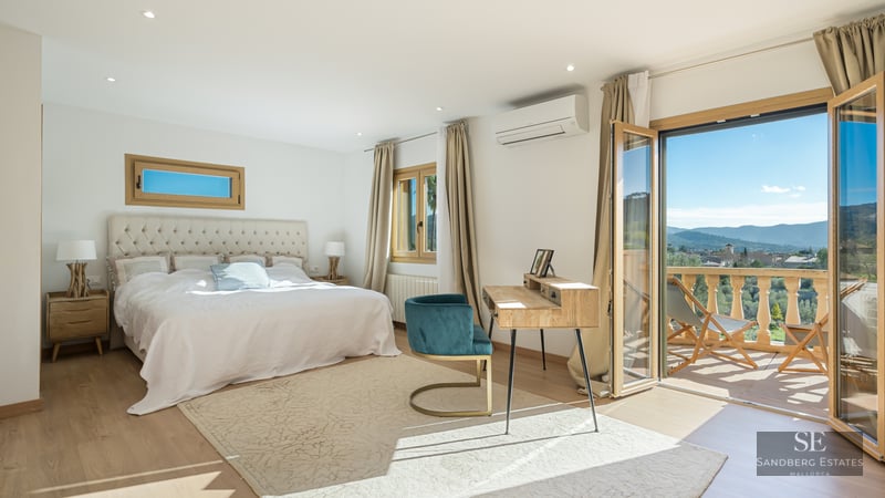 Bright master bedroom with a large bed, wooden desk, teal chair, and open glass doors leading to a scenic balcony.