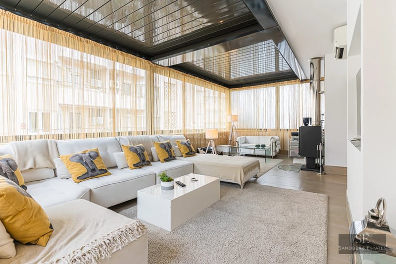 Spacious, bright living room with large windows, light wood floors, and minimalist decor. Beige sofa, coffee table, and access to terrace.