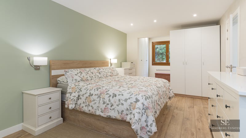 Modern bedroom featuring a floral bed, sage green wall, white furniture, and light wood flooring.
