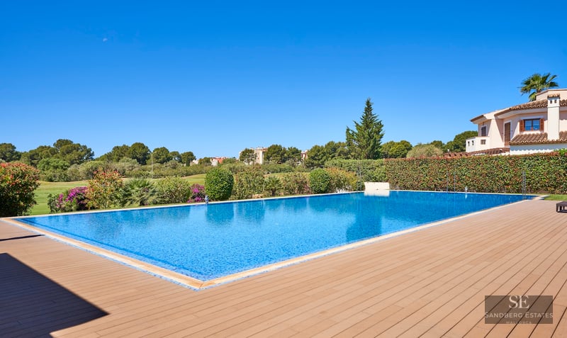 Large blue swimming pool with a wooden deck, surrounded by green hedges and a Mediterranean villa under a clear sky.