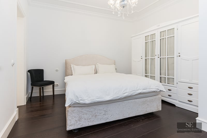 Bright bedroom featuring a beige bed, large white wardrobe, black chair, and crystal chandelier on dark wood floors.