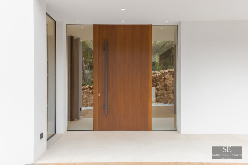 Large wooden pivot door with glass side panels flanked by smooth white walls and minimalist lighting.