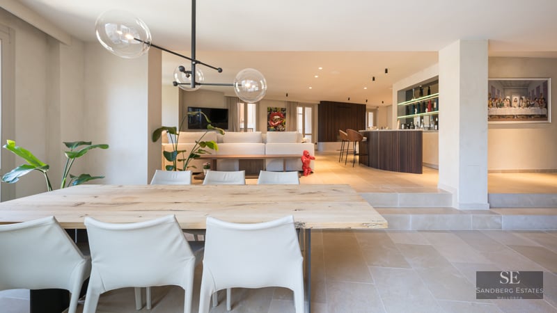 A spacious modern open-plan room featuring a wooden dining table, white chairs, glass globe pendants, and a bar area.