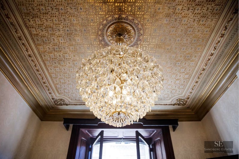 A large crystal chandelier hanging from a highly decorative gold-patterned ceiling with ornate moldings.