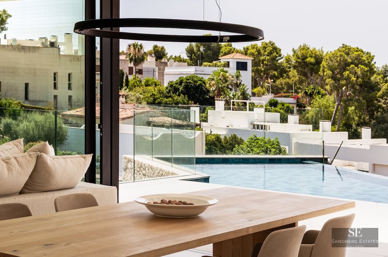 A minimalist dining room with a wooden table and glass walls overlooking a bright blue infinity pool and greenery.