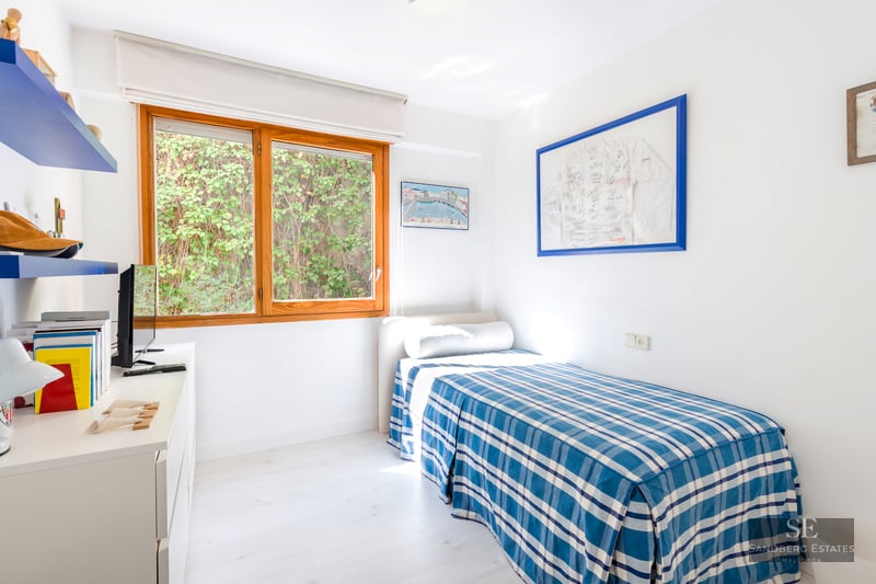 Bright single bedroom featuring a blue checkered bed, white desk, and a large wooden window looking onto green foliage.