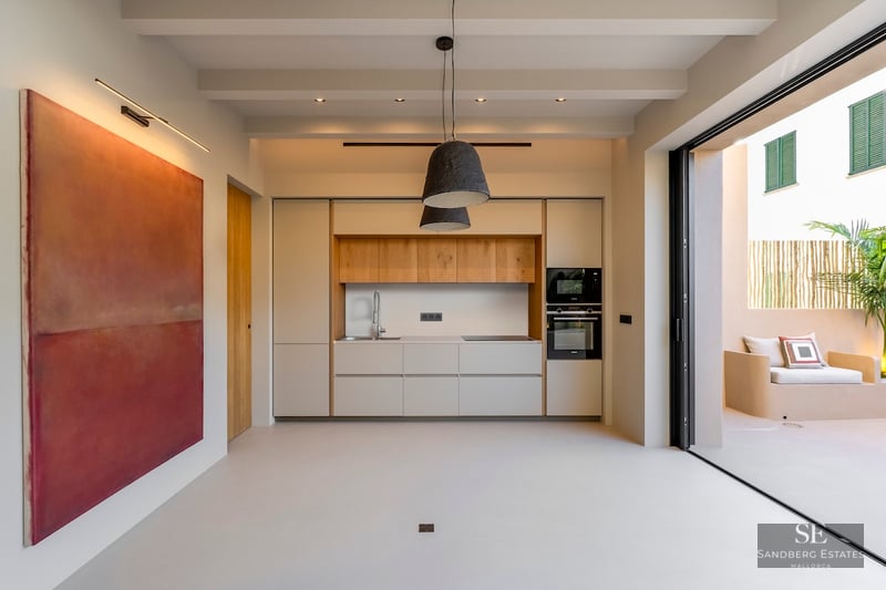 Cuisine minimaliste avec accents en bois clair, grand tableau rouge et baies vitrées ouvrant sur un patio ensoleillé.