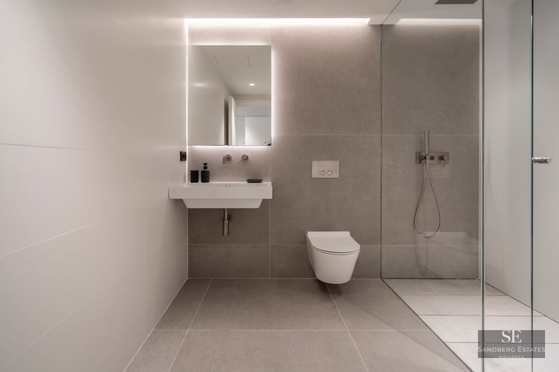 Modern bathroom featuring grey tiles, a backlit mirror above a white sink, a wall-hung toilet, and a glass shower partition.