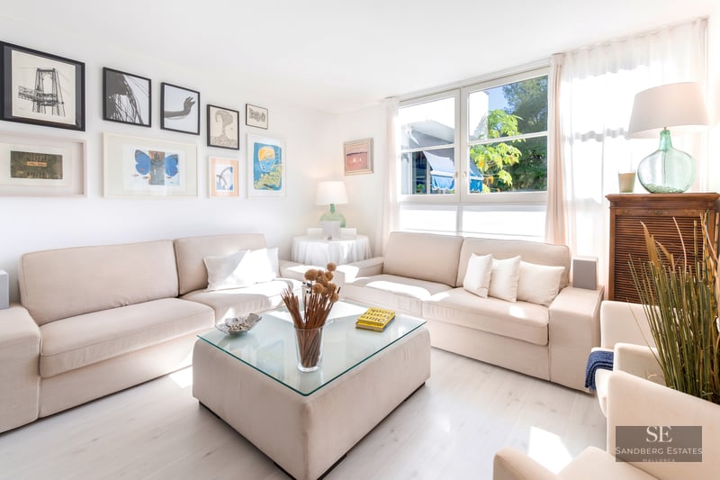 Bright living room with two beige sofas, a glass-top coffee table, large windows, and a wall of framed art.