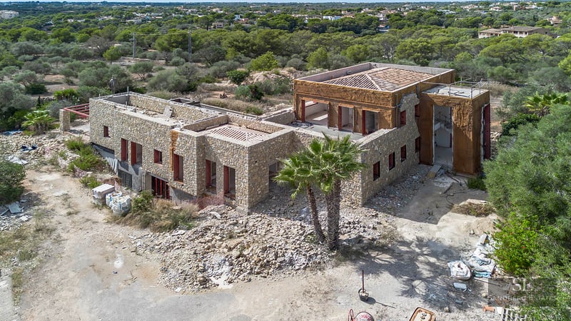 Elevated view of a large stone-walled luxury villa currently under construction surrounded by Mediterranean vegetation.