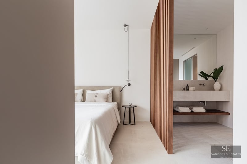 Modern bedroom with a beige bed and a wooden slat divider separating a stone vanity bathroom area.