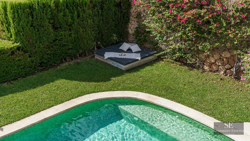 A green-tiled swimming pool next to a manicured lawn and a daybed with pillows under pink bougainvillea flowers.