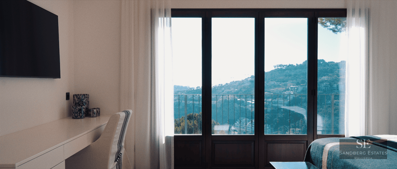 A modern bedroom featuring a desk, a wall-mounted TV, and floor-to-ceiling windows overlooking a scenic hillside and sea.
