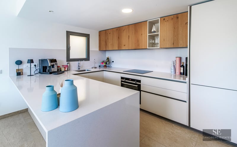 Modern white kitchen with wooden upper cabinets, a large white island, and built-in appliances.
