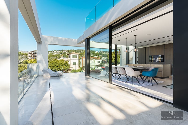 Spacious white terrace with glass railings looking into a modern dining room through large sliding glass doors.