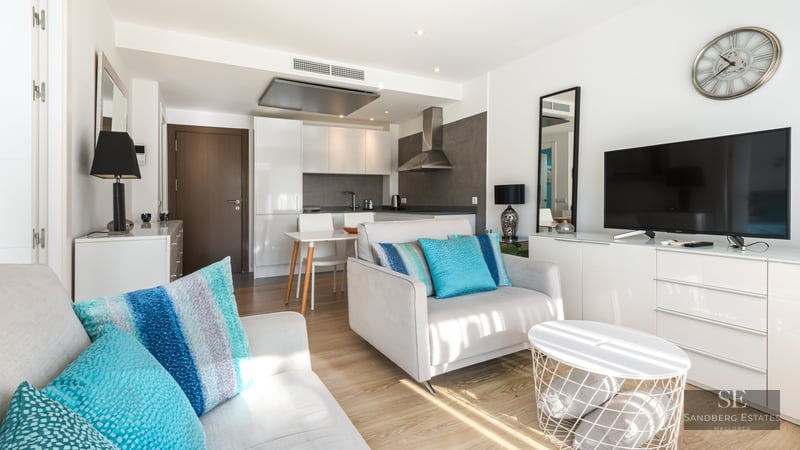 Salon lumineux avec fauteuil gris, coussins turquoise, meuble TV et cuisine moderne en arrière-plan.