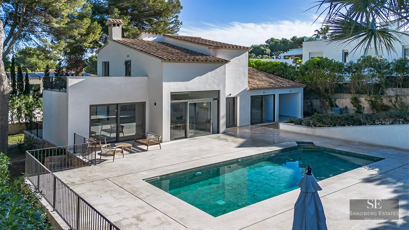 White Mediterranean-style villa with a rectangular blue swimming pool and a large stone deck under bright sunlight.