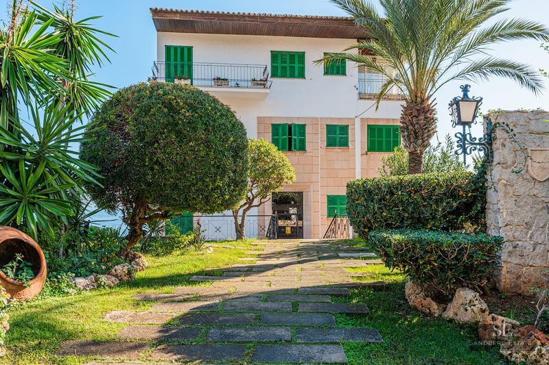 Stone pathway leads through a lush garden with palm trees to a white villa with vibrant green shutters.