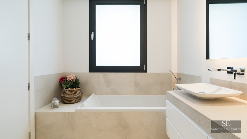 Minimalist bathroom featuring a white built-in tub, vessel sink on a stone vanity, and a black-framed window.