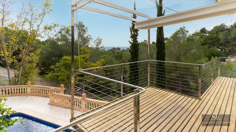 Elevated wooden deck with metal railings overlooking a pool and lush green forest under a blue sky.
