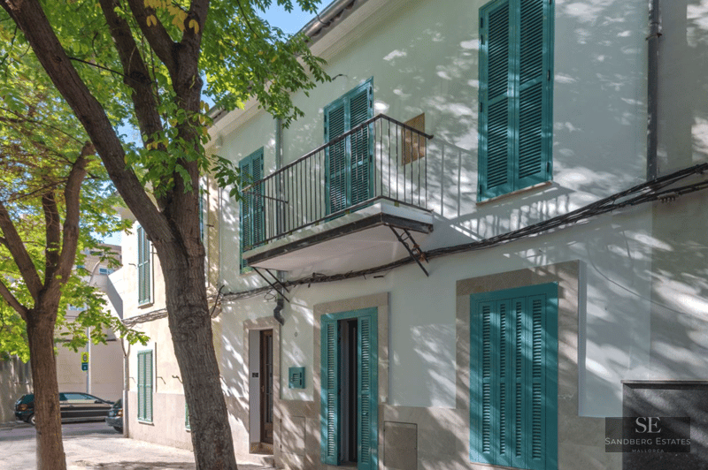 White building exterior featuring turquoise shutters, a small iron balcony, and shadows from a large leafy tree.