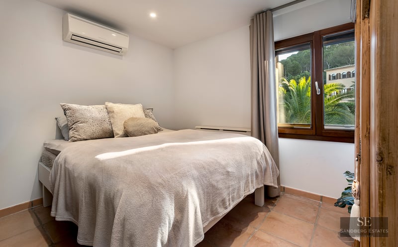 Bedroom with a cozy double bed, terracotta floors, air conditioning, and a window overlooking lush green palm trees.