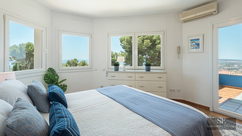 Bright white bedroom with large windows overlooking the sea, blue bedding, and a door to a sunny balcony.