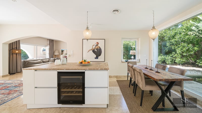 A modern white kitchen island with a wine fridge next to a rustic wooden dining table overlooking a lush garden.