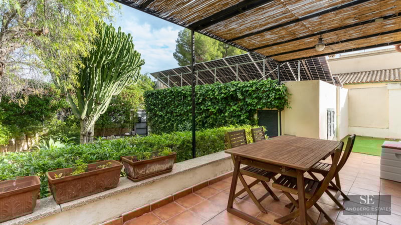 Wooden dining table under a bamboo pergola on a terracotta terrace surrounded by lush greenery and solar panels.