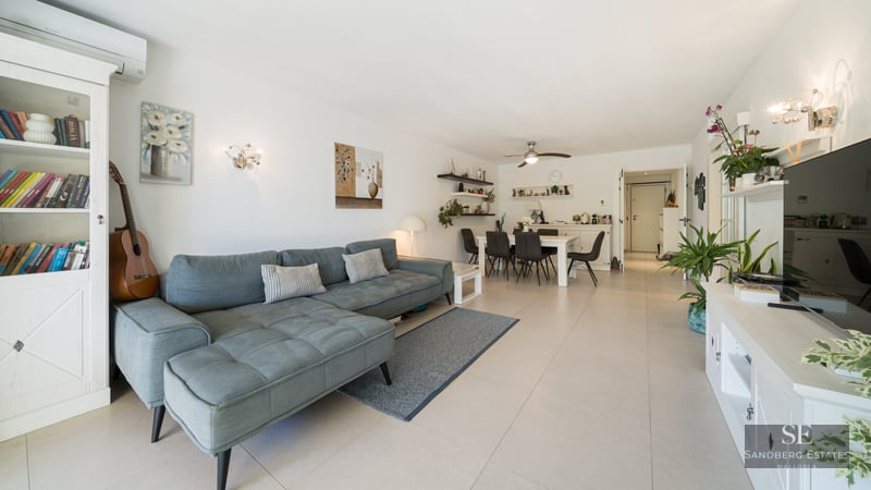 Salon lumineux avec un grand canapé en L bleu, des meubles blancs, un coin repas et un sol carrelé.