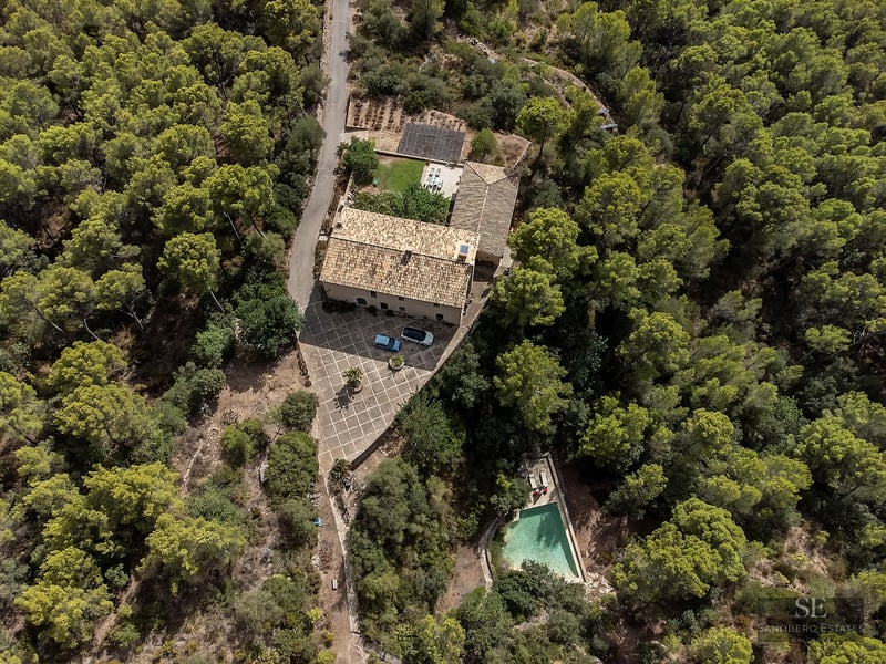 Aerial view of a stone house with terracotta roof, courtyard, and a pool, all surrounded by a dense green pine forest.