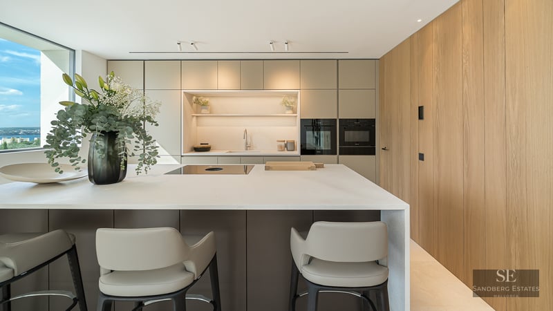 Modern kitchen island with stools, built-in appliances, wooden wall, and large window overlooking the water.