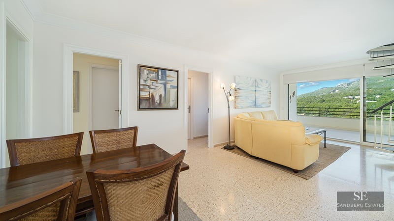 Open living space with a cream leather sofa, wooden dining set, and large glass doors revealing a mountain landscape.