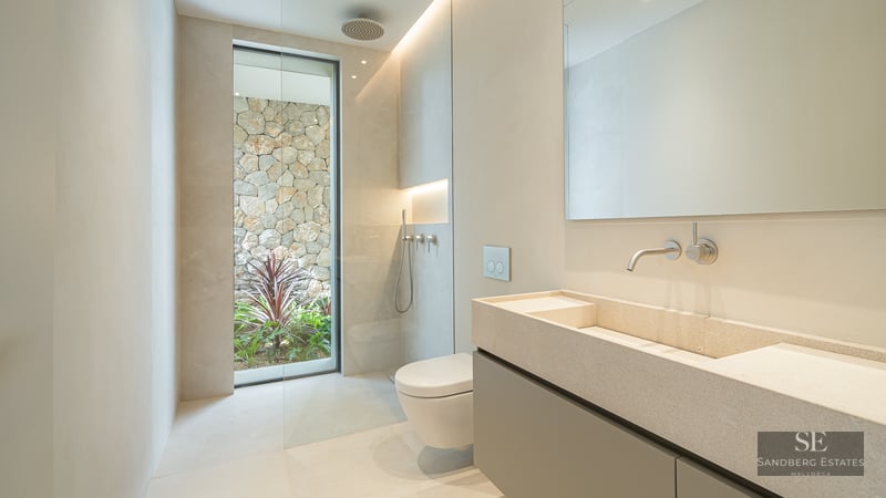 Minimalist bathroom with a stone double vanity, walk-in shower, and floor-to-ceiling window facing a natural stone wall.