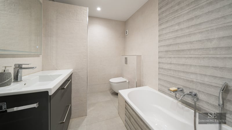 Salle de bain moderne avec baignoire blanche, WC suspendu et meuble gris sur fond de carrelage beige texturé.